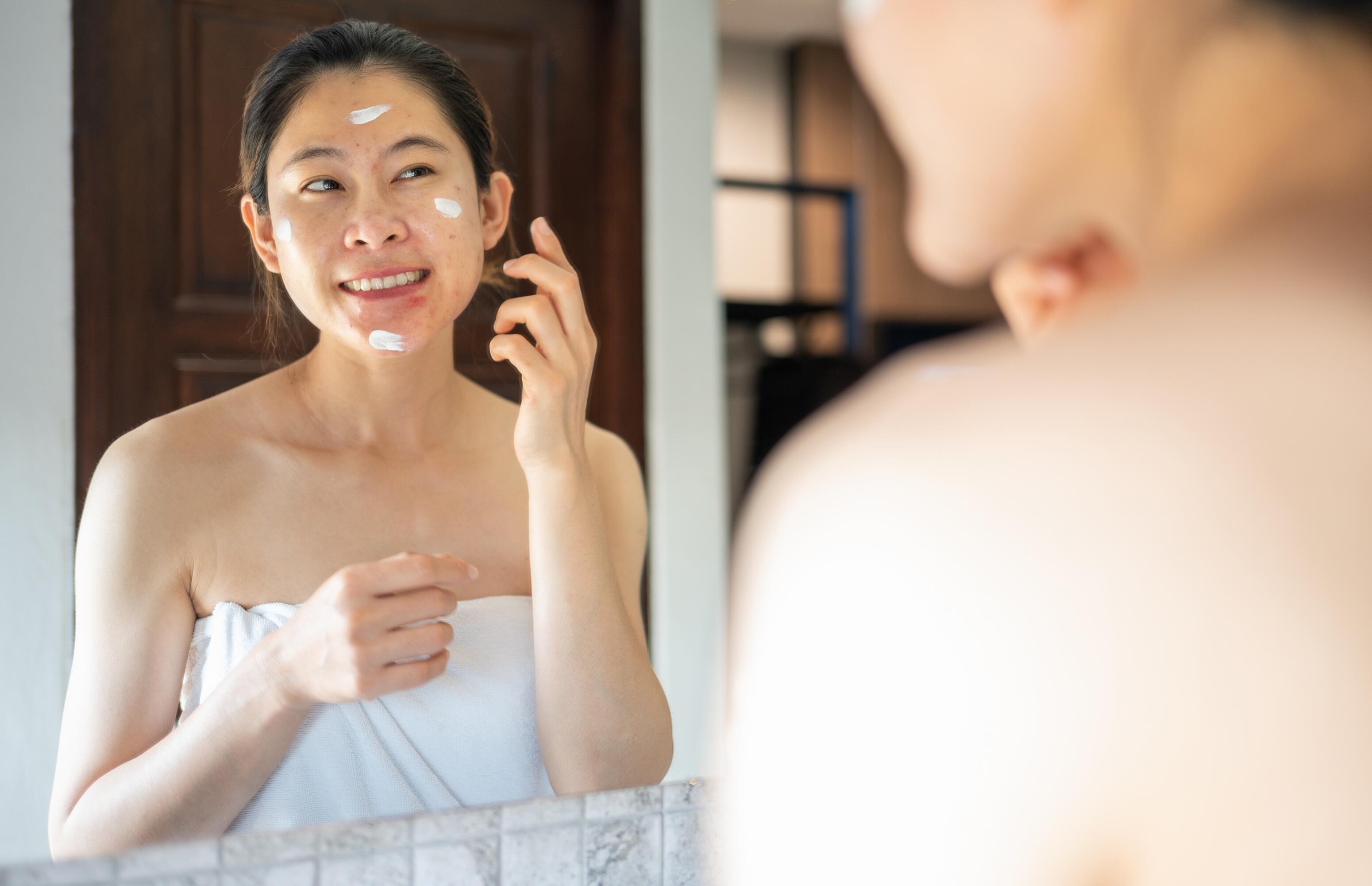 Eine asiatische Frau in einem weißen Handtuch schaut lächelnd in den Spiegel, sie trägt weiße Creme auf ihrem Gesicht auf.